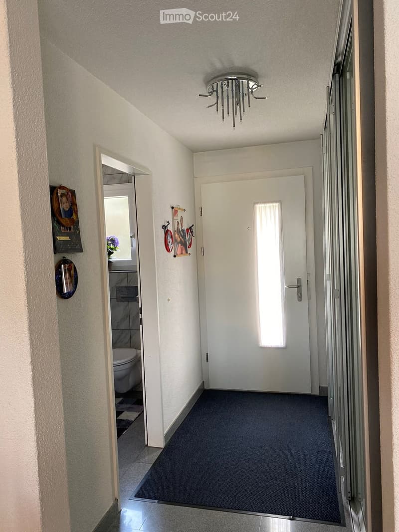 A narrow hallway with a carpeted floor, white walls, and a ceiling light fixture. There are two doors, one leading to a bathroom and the other to another room. The hallway features a small decorative mirror and a calendar on the wall.