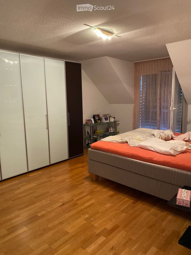 Bedroom with wooden flooring, a grey bed with orange bedding, a white wardrobe, a small shelf with decorative items, and a nightstand with a gift box.