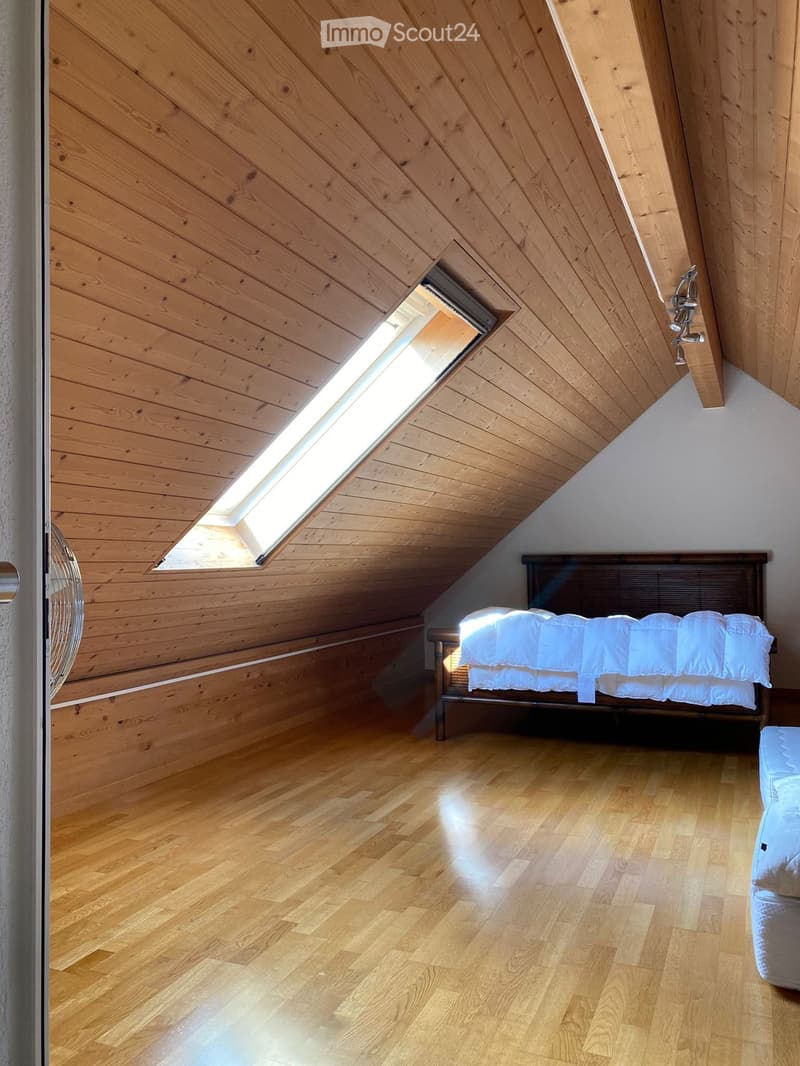 Bright attic bedroom with wooden walls and floor, skylight window, bed with white bedding, and a fan on the left side.