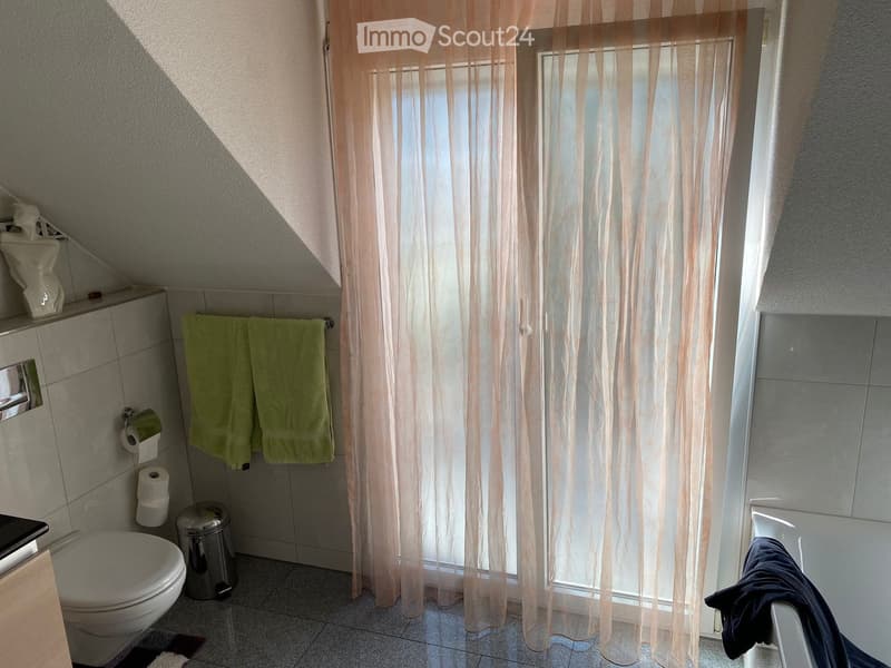 Bathroom with a toilet, bathtub, green towels, toilet paper, and a trash can. Light-colored curtains cover the window, and a small shelf holds decorative items.