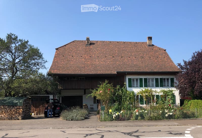 Two-story house with a red-tiled roof, white walls, green shutters, and a wooden balcony. Features a garage, garden with flowers, and a stone woodpile.