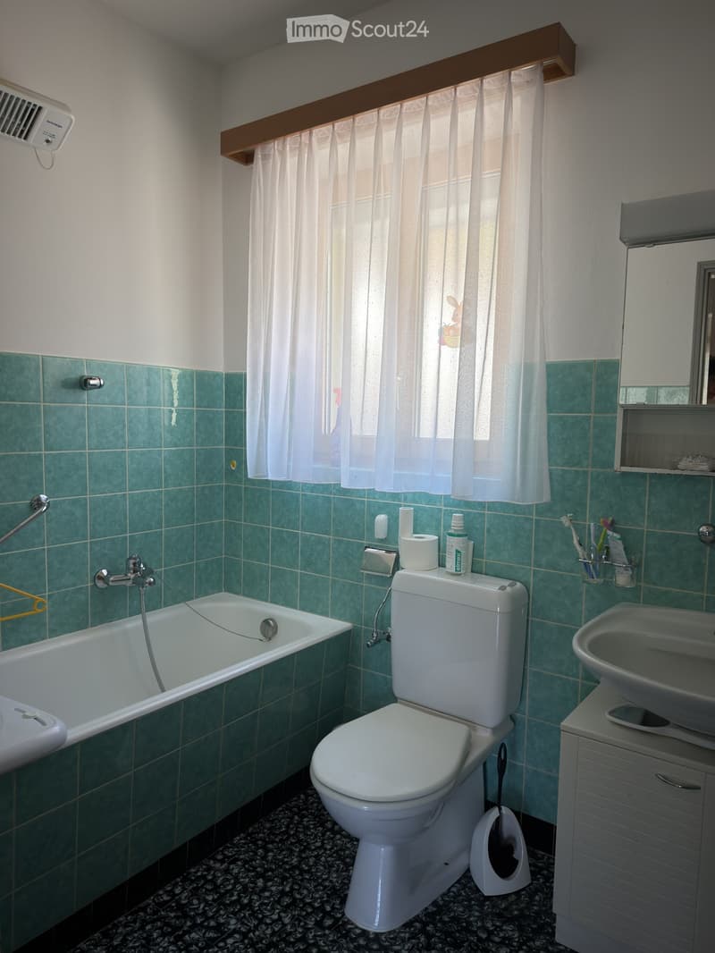 Bathroom with green tiled walls, white bathtub, toilet, sink, mirror, and cabinet. Toilet paper and toothbrushes are visible on the sink counter.