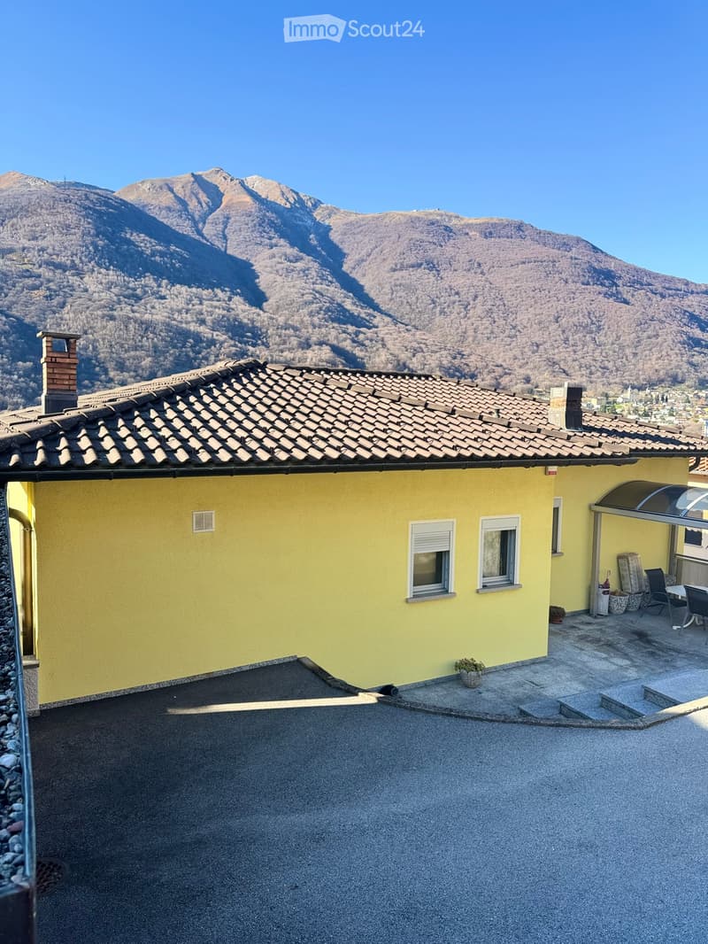 Yellow house with tiled roof, chimney, and windows, featuring a small outdoor seating area with a table and chairs, overlooking a mountainous landscape.