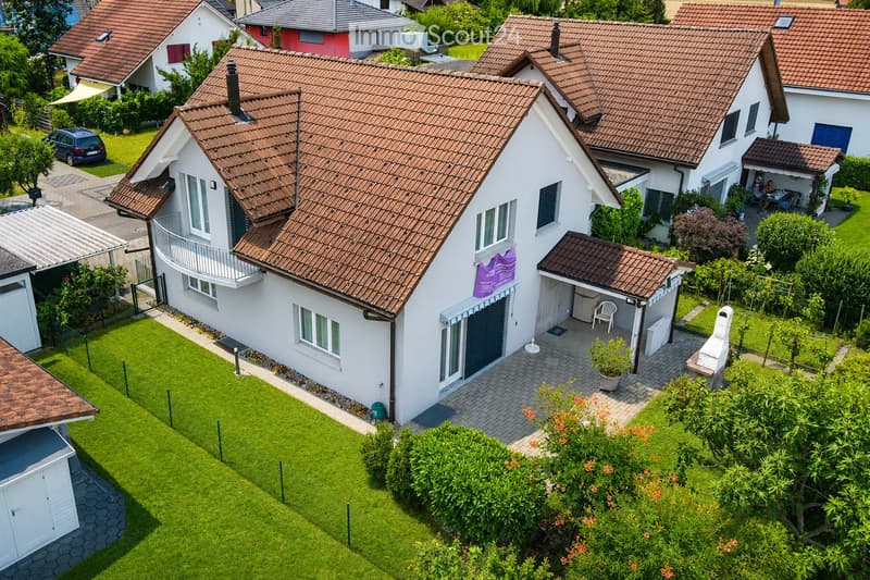A two-story house with a red-tiled roof, white exterior, and a well-maintained garden with a barbecue and seating area.