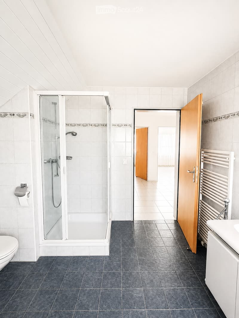 Modern bathroom with white tiles, dark grey floor tiles, a glass-enclosed shower, toilet, sink, and radiator.
