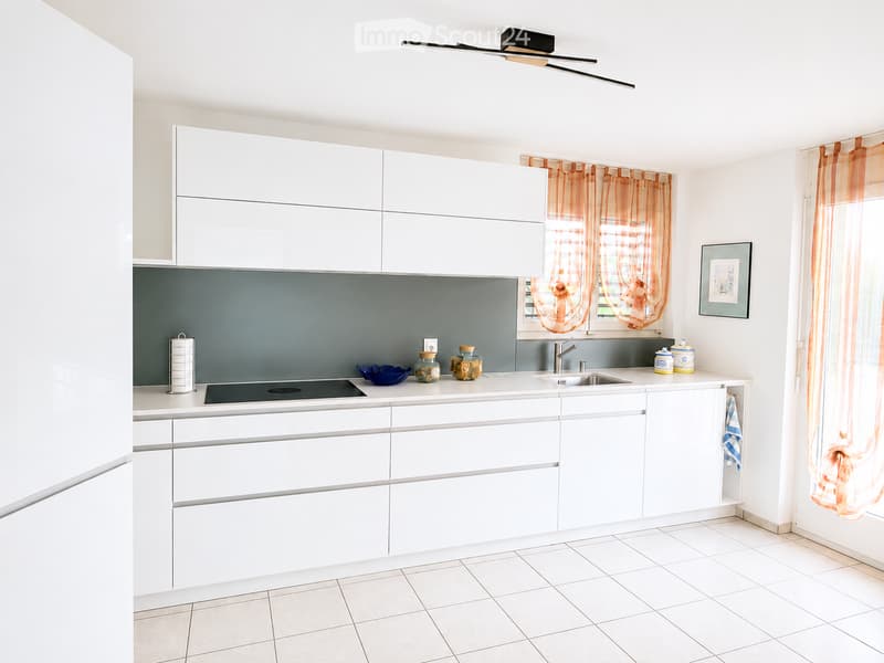 Modern kitchen with white cabinets, a grey backsplash, and a tiled floor. Features a sink, faucet, and various small decorative items on the counter.