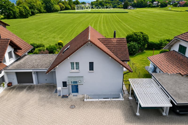 A modern two-story house with a red-tiled roof, light gray exterior, and a spacious driveway. The house features a garage, multiple windows, and is surrounded by a well-maintained lawn and trees.
