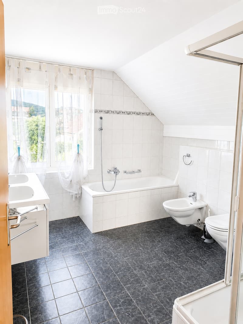 Modern bathroom with white tiles, dark grey marble flooring, a bathtub, double sink, toilet, bidet, and a showerhead. A towel hangs on the side.
