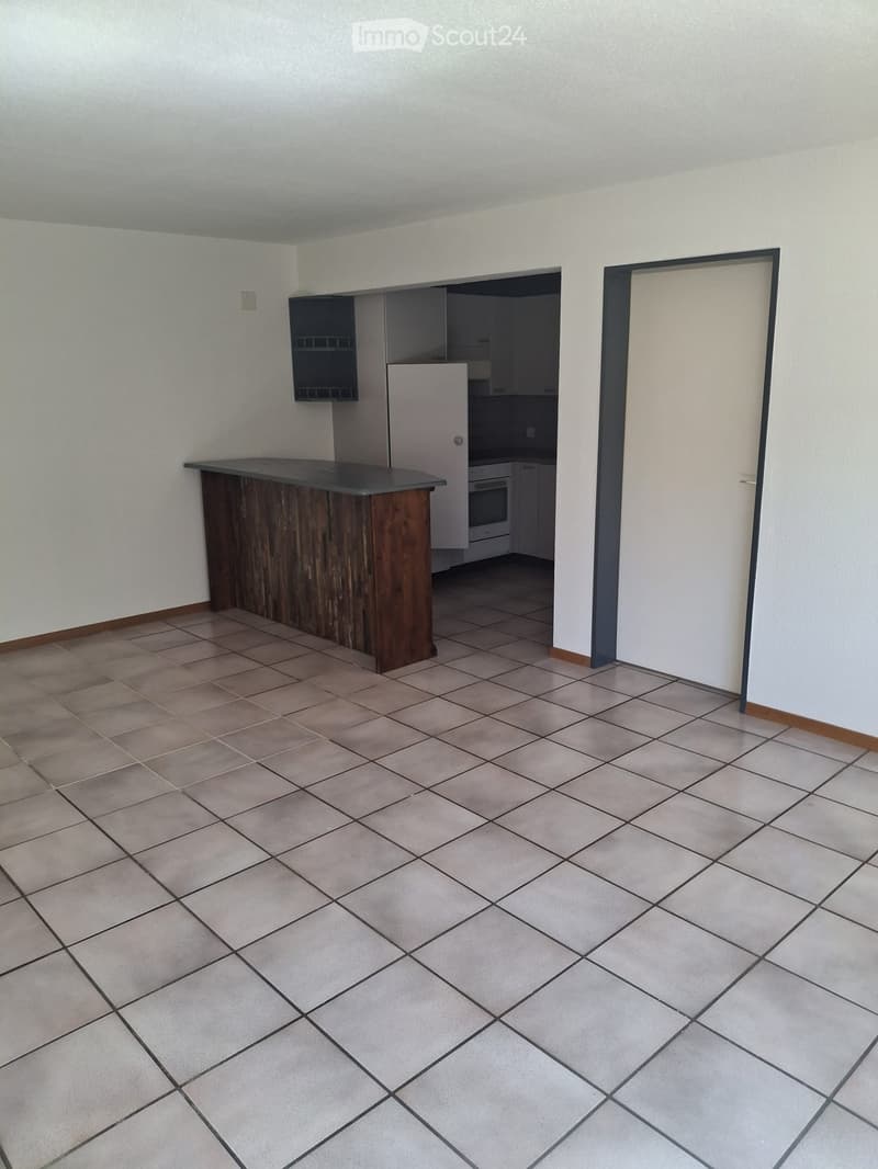 Empty room with tiled floor, white walls, and a wooden counter. Kitchen area visible in the background with cabinets and appliances.