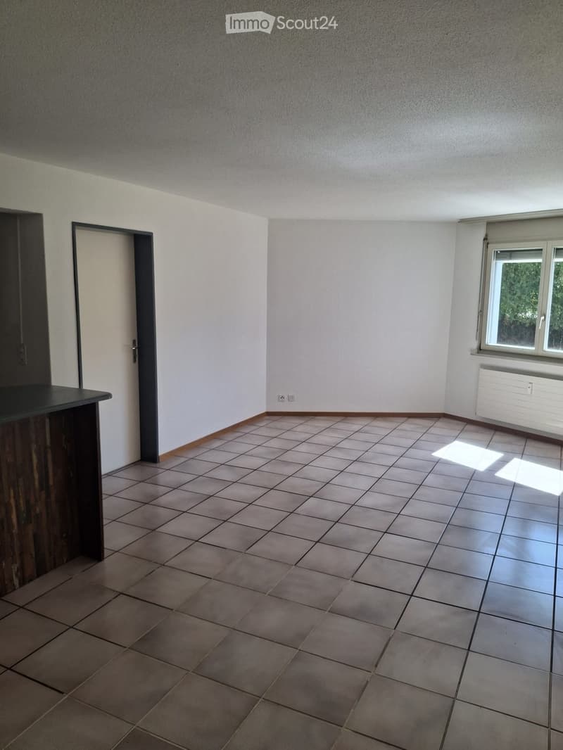 Empty room with tiled floor, white walls, and a window with blinds. There is a door on the left side and a radiator below the window.