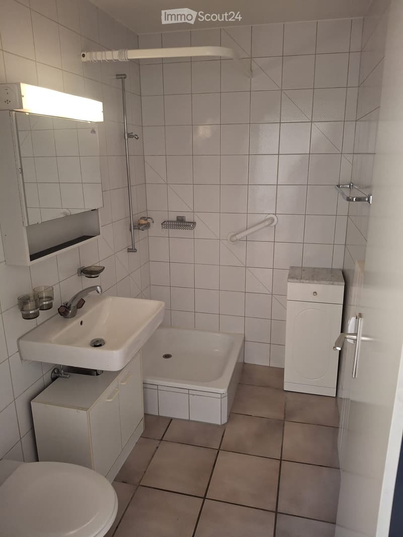 Bathroom with a white sink, bathtub, toilet, mirror, and cabinet. Features tiled walls and floor, a handrail, and a small shelf with glasses.
