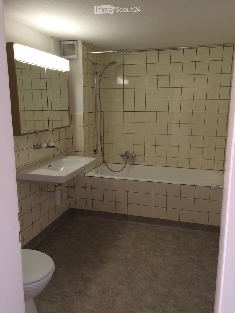 Bathroom with tiled walls, a toilet, a sink, a bathtub, and a mirror with a built-in light. The floor is concrete.