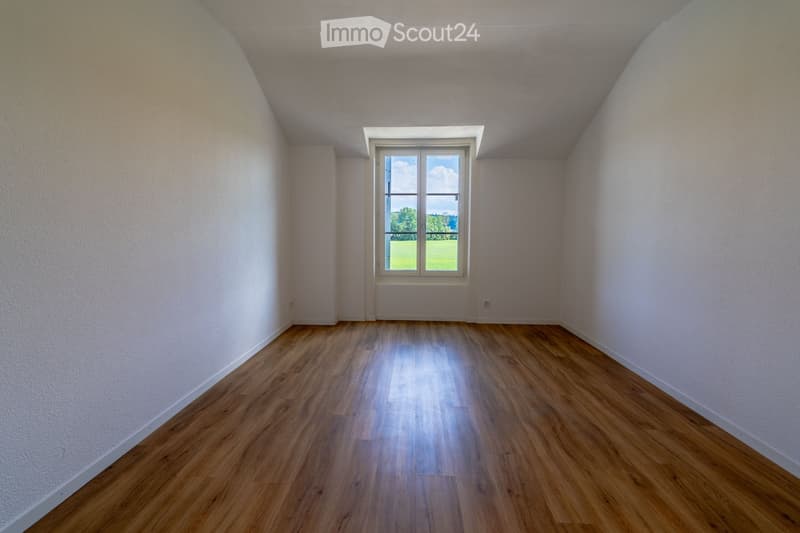 Empty room with wooden flooring and white walls, featuring a large window with a view of a green landscape.