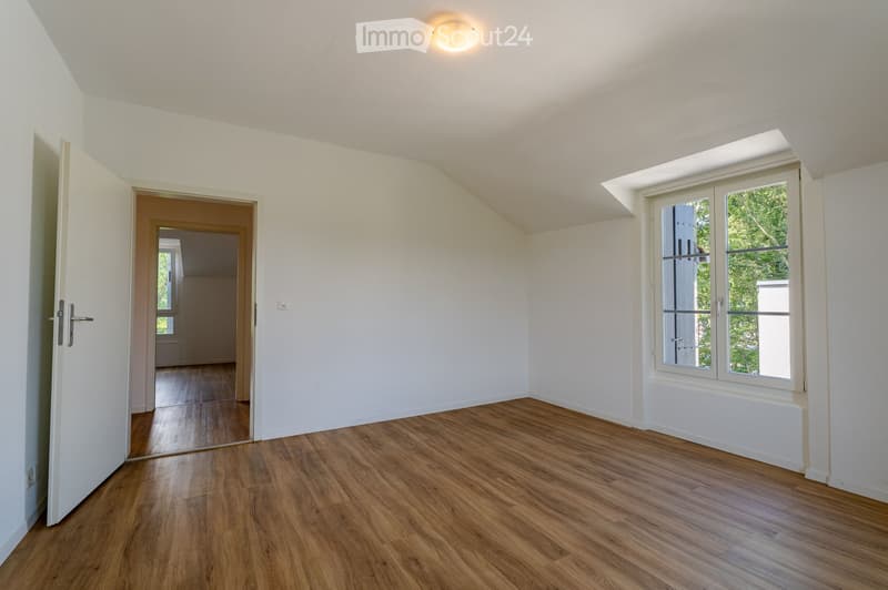 Empty room with wooden flooring, white walls, and a large window with a view of greenery. The room features a door leading to another space and a ceiling light fixture.