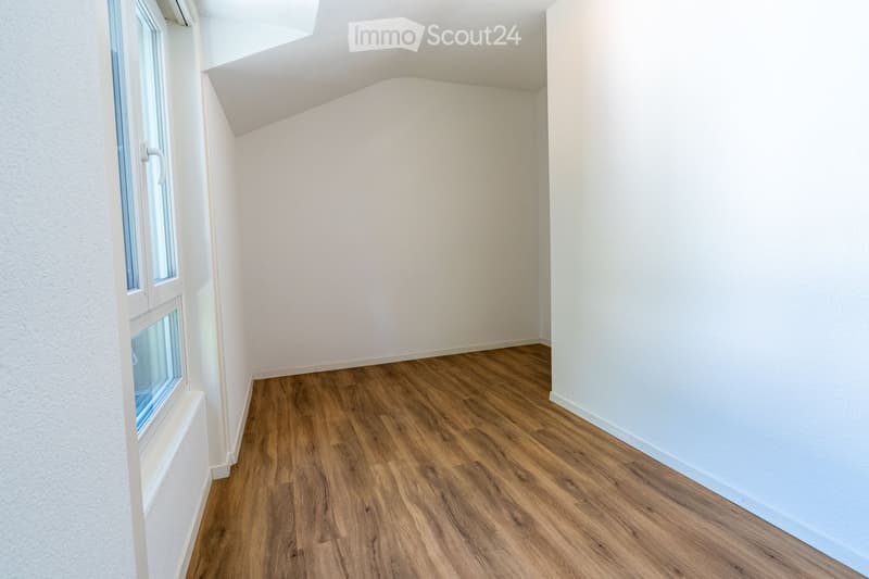 Empty room with wooden flooring and white walls, featuring a large window with white frames and a simple, sloped ceiling.