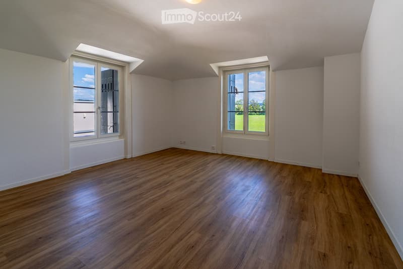 Empty room with wooden flooring, white walls, and large windows offering a view of the outdoors.