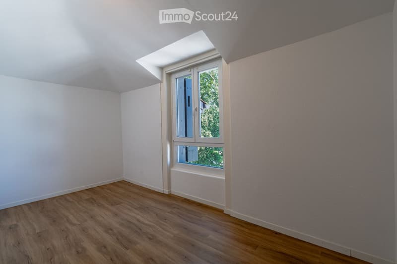 Empty room with wooden flooring, white walls, and a large window with a view of greenery outside.