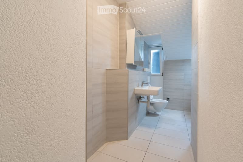 Modern bathroom with white tiles, a sink, toilet, mirror, and cabinet. The space is clean and well-lit with a window providing natural light.