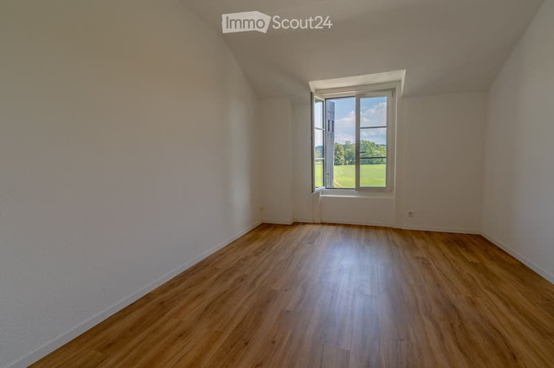Empty room with wooden flooring, white walls, and a large window offering a view of a green landscape.