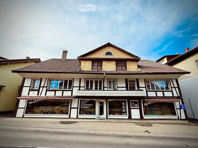 Two-story building with a tiled roof, white and brown facade, large windows, and a balcony on the upper floor. The ground floor has storefronts with glass windows and awnings.
