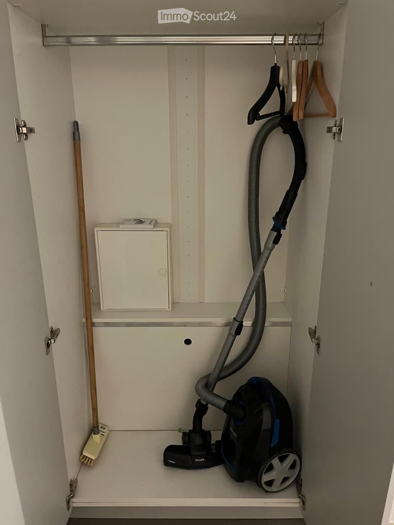 Open closet with white walls and shelves, containing a vacuum cleaner, broom, and hangers.