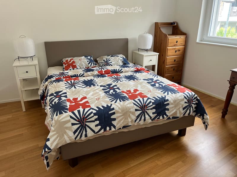Bedroom with a wooden floor, a large bed with a colorful quilt, two nightstands with lamps, and a wooden dresser with drawers.