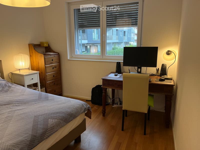 Cozy bedroom with a bed, desk, chair, and a view of the city through large windows. Includes a lamp, drawer unit, computer monitor, and speaker.