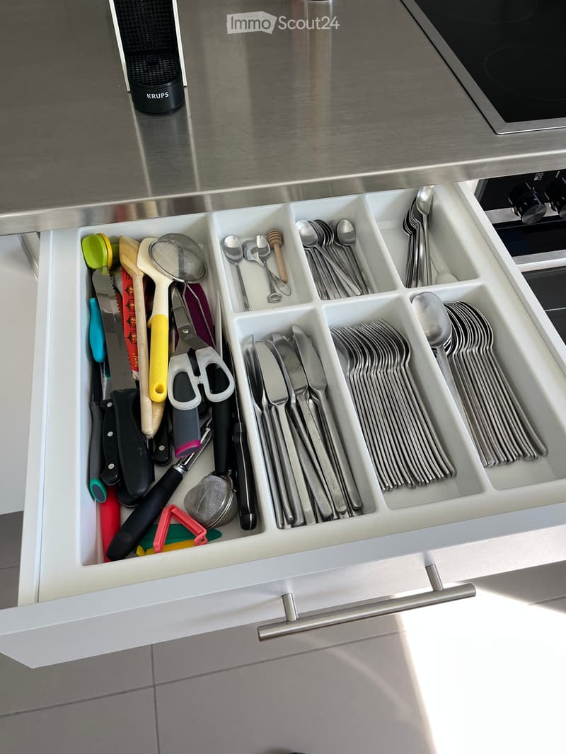 Open kitchen drawer with neatly organized cutlery and utensils, including knives, forks, spoons, measuring spoons, a strainer, and scissors.