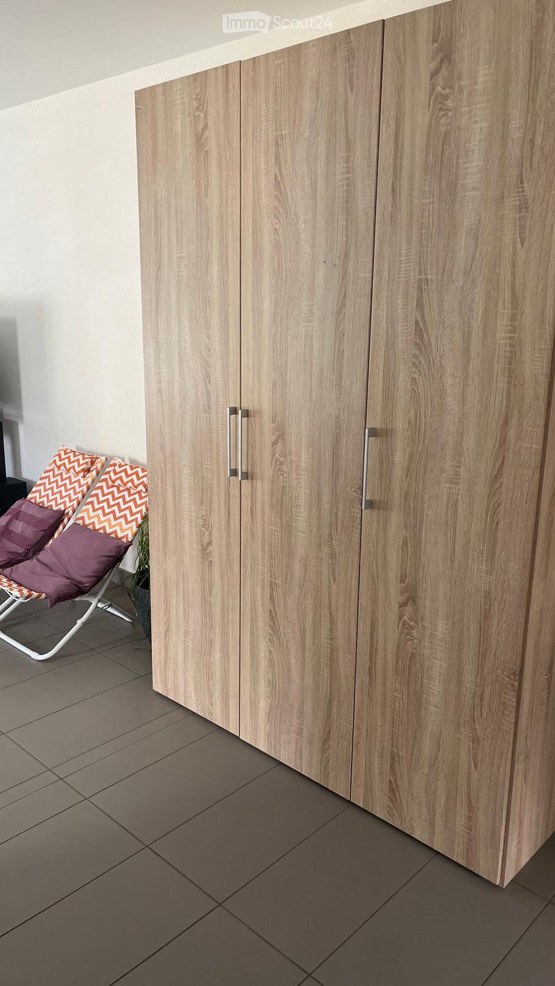 A corner of a room with light wooden cabinets and a grey tiled floor. A white folding chair with orange and purple cushions is placed near the cabinets.