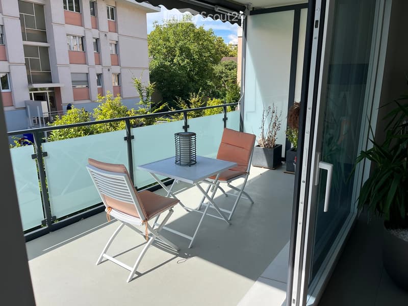 Balcony with a small table, two chairs, a candle holder, and potted plants, overlooking a residential area with trees.