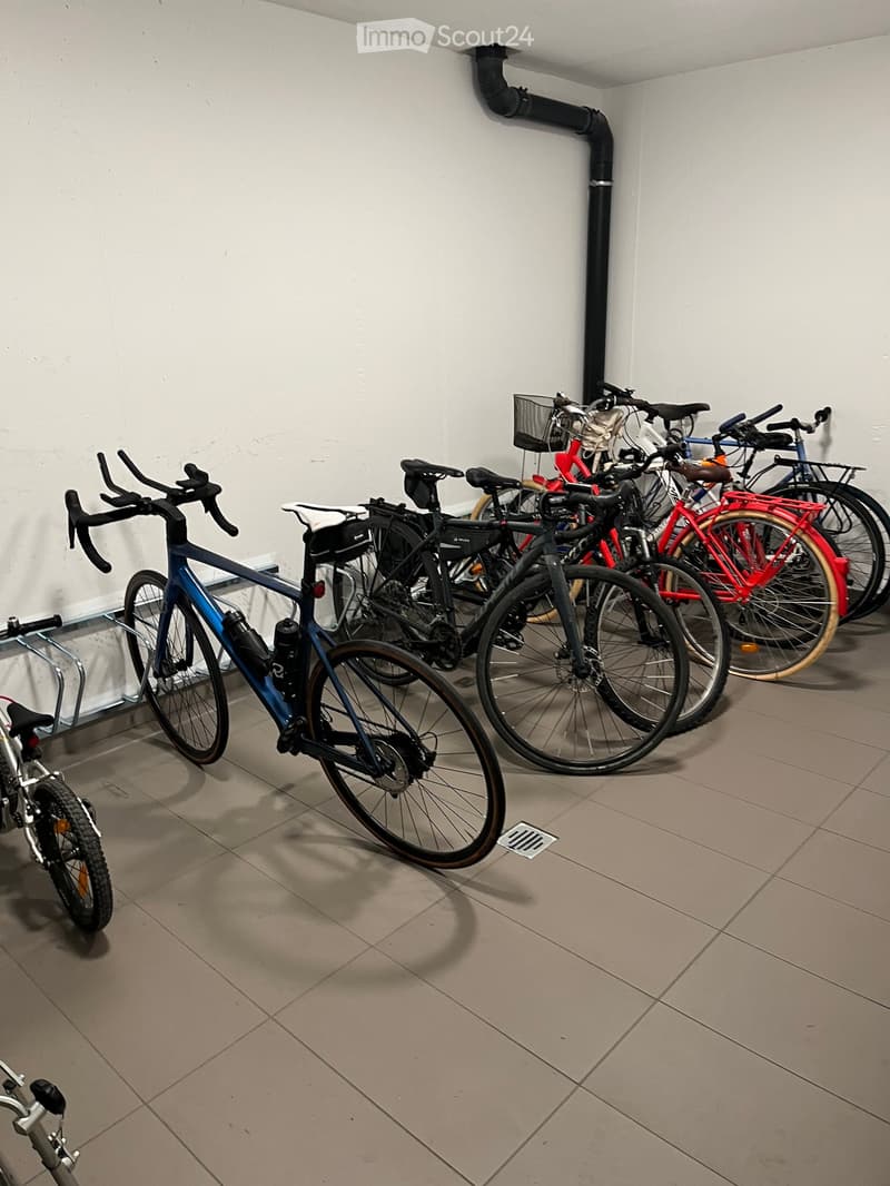 A storage area with several bicycles parked in a row against a white wall, featuring tiled flooring and a visible ventilation pipe.