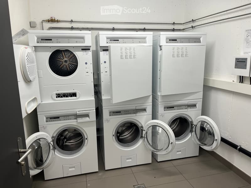 Three stacked washing machines and dryers in a clean, tiled room with white walls and exposed pipes.