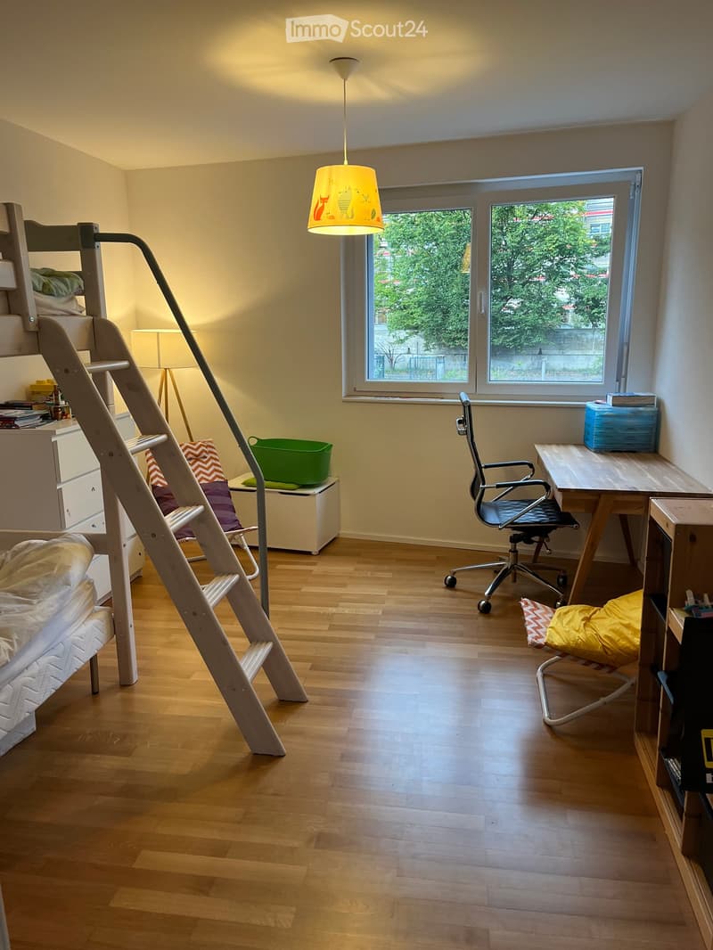A well-lit bedroom with wooden flooring, a bunk bed, a desk with a chair, a hanging lamp, and a small storage unit.