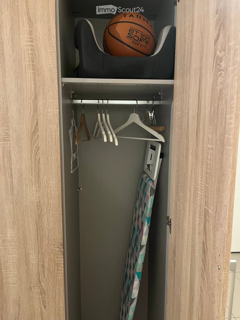 Open closet with wooden doors, metal rod with hangers, and a shelf holding a sports ball and folded items.