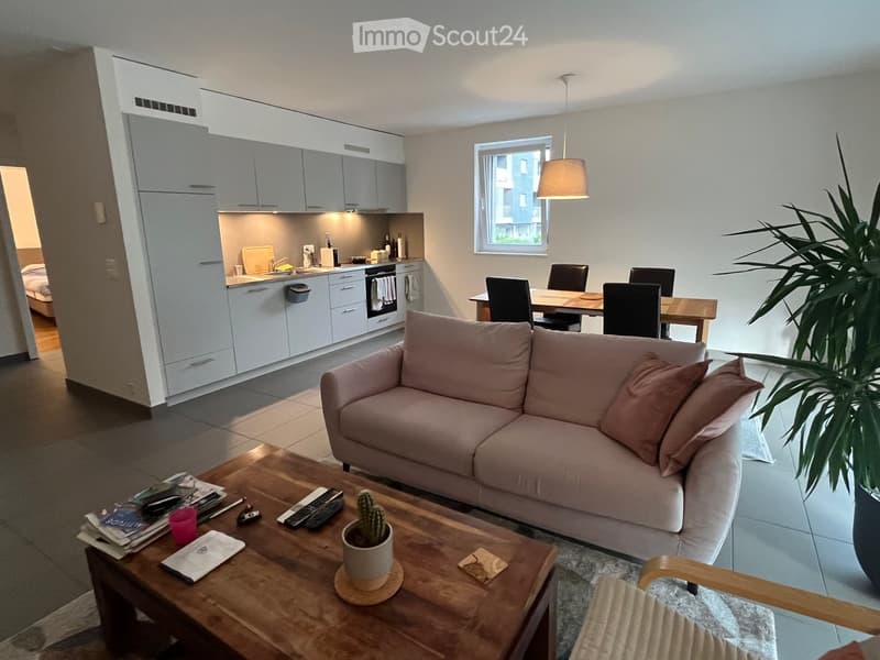 A modern, open-concept living and dining area with a light pink sofa, wooden coffee table, dining table with chairs, and a small kitchen area with white cabinets and stainless steel appliances.