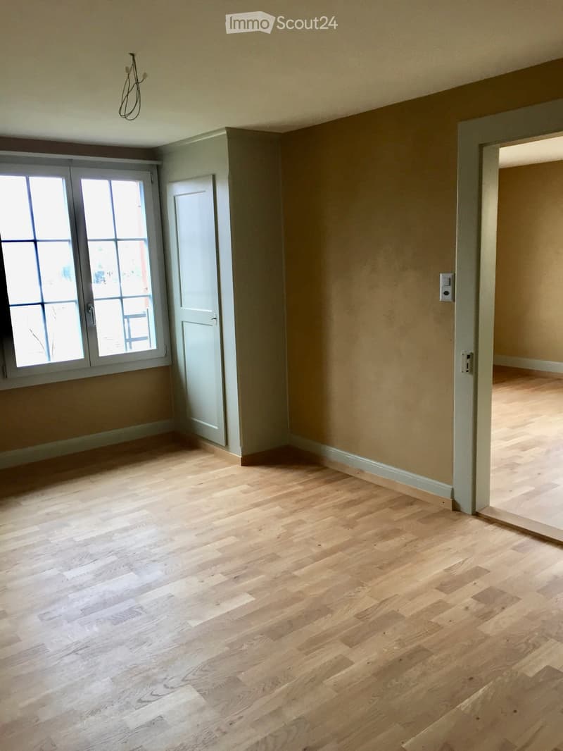Empty room with light wooden flooring, beige walls, and a large window with glass panels. A white door is visible on the left side, and a light switch is on the right wall.