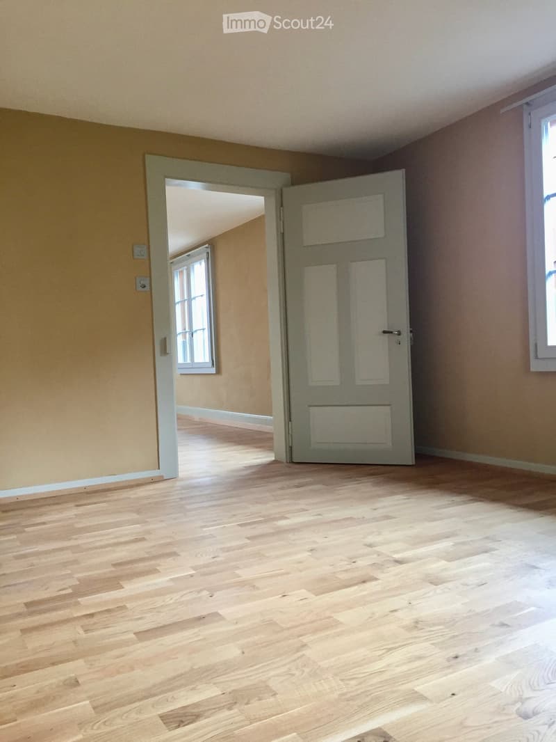 Empty room with light wooden flooring, light-colored walls, and an open door leading to another room with similar flooring and walls.