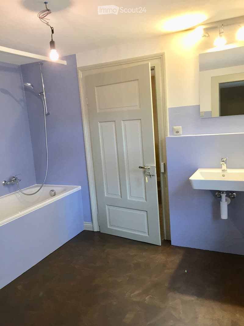 Bathroom with a white bathtub, a white sink, a mirror, and a door leading to another room. The walls are painted in a light blue color, and the floor is dark brown.