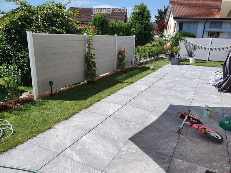 Well-maintained garden with a paved patio, green lawn, and surrounding houses. Features include a white fence, potted plants, a small bicycle, and a garden hose.
