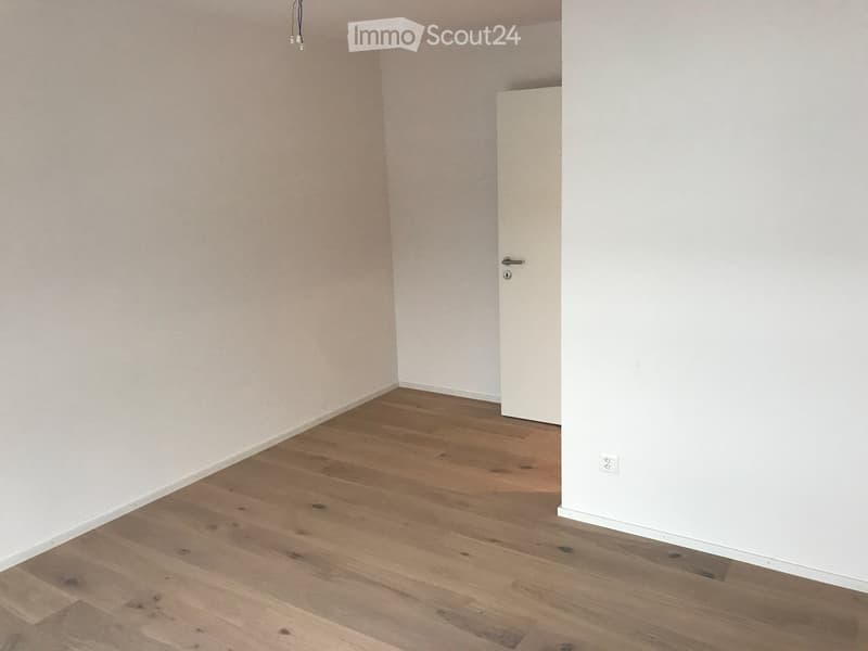 Empty room with light wooden flooring and white walls, featuring a closed white door and a visible electrical outlet.