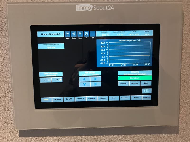 A wall-mounted control panel with a digital screen displaying weather and home automation controls, including temperature, lighting, and heating settings.