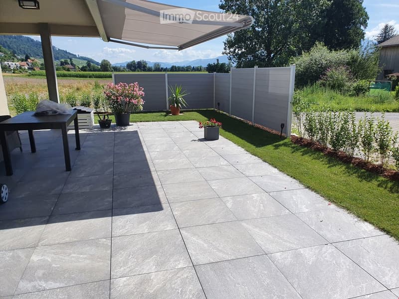 Outdoor patio with tiled floor, table, chairs, and potted plants, surrounded by a white fence and green lawn.