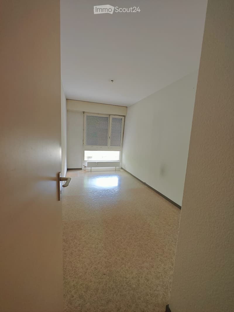 Empty hallway with beige walls, carpeted floor, and a window with blinds at the end.