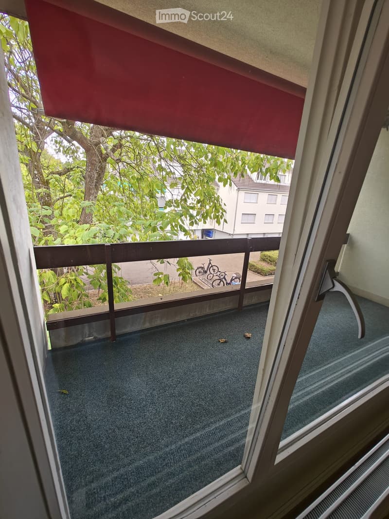 Balcony with carpeted floor, wooden railing, and a view of greenery and neighboring buildings.