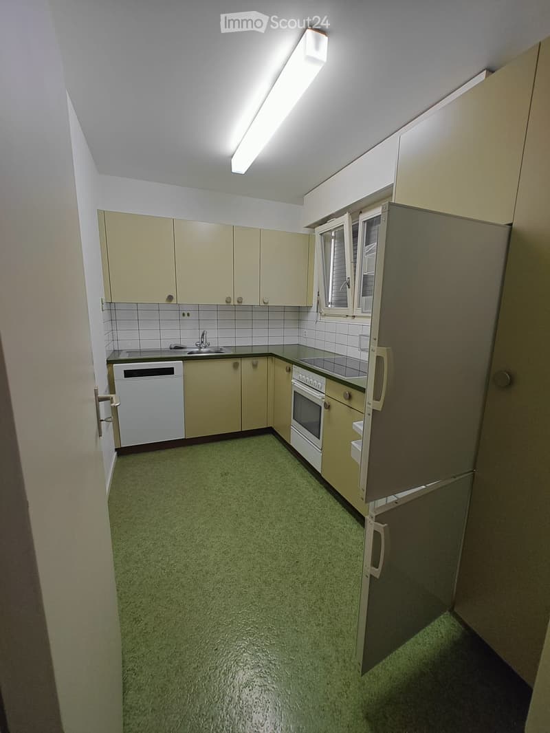 Empty kitchen with light-colored cabinets, a refrigerator, oven, dishwasher, and a sink, featuring a green floor and white tiled backsplash.