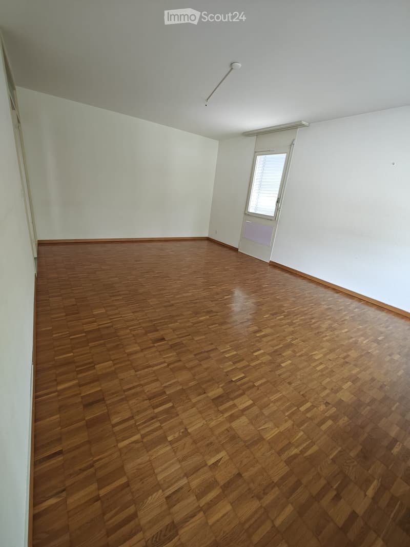 Empty room with wooden parquet flooring and white walls, featuring a single window with blinds and a door.