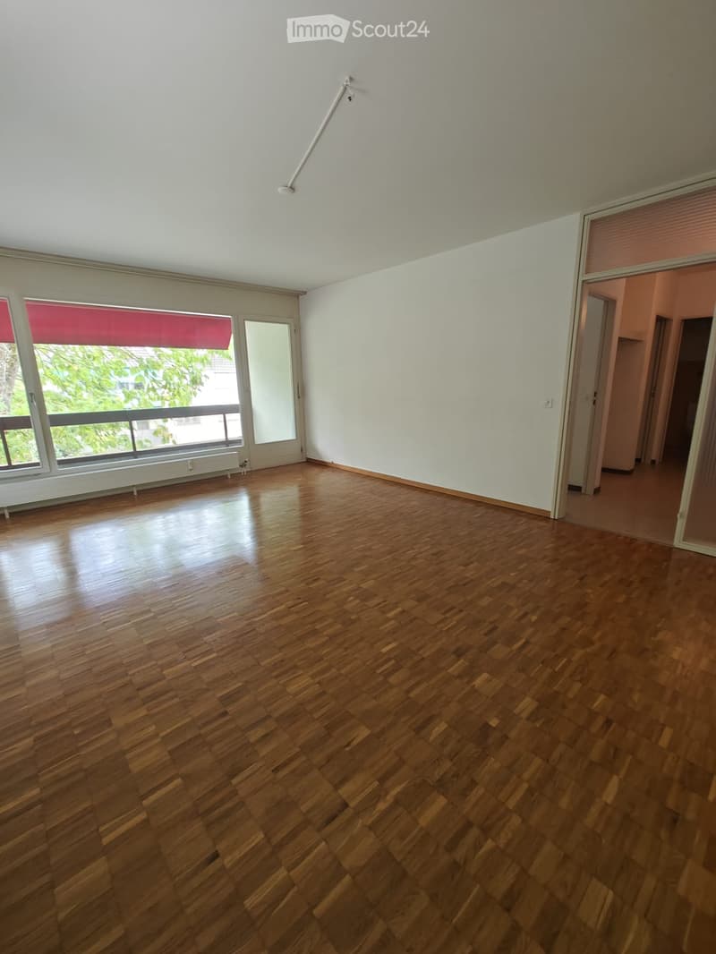 Empty room with wooden flooring, large windows with red blinds, and a visible hallway leading to another area.