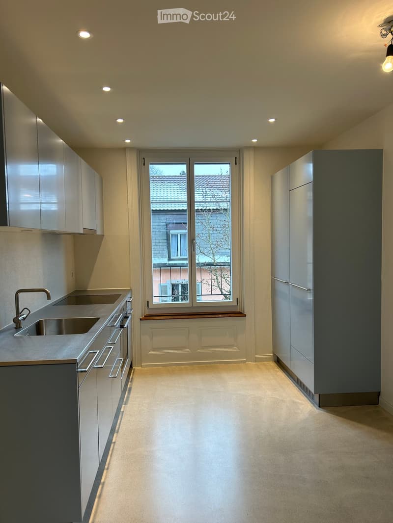 Modern, minimalist kitchen with sleek white cabinets, stainless steel sink, and a large window overlooking a neighboring building. The space is clean and uncluttered, with a smooth concrete floor and recessed lighting.