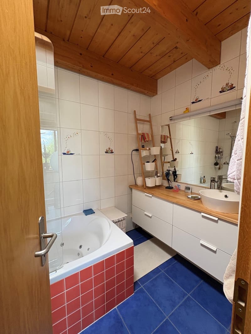 Bathroom with a wooden ceiling, white tiles, a bathtub with red tiles, a sink with a wooden countertop, a mirror, a ladder with towels, and a toilet.