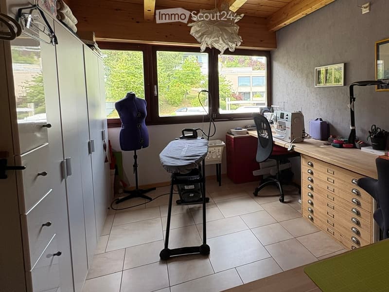 Well-lit room with wooden ceiling beams, large windows, and tiled floor. Contains a wardrobe, desk with sewing machine, ironing board, mannequin, and a chair.